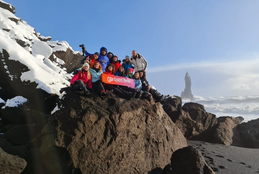 The group posing with the Sto Gran Tour banner on a snowy rock, in front of an Icelandic black sand beach