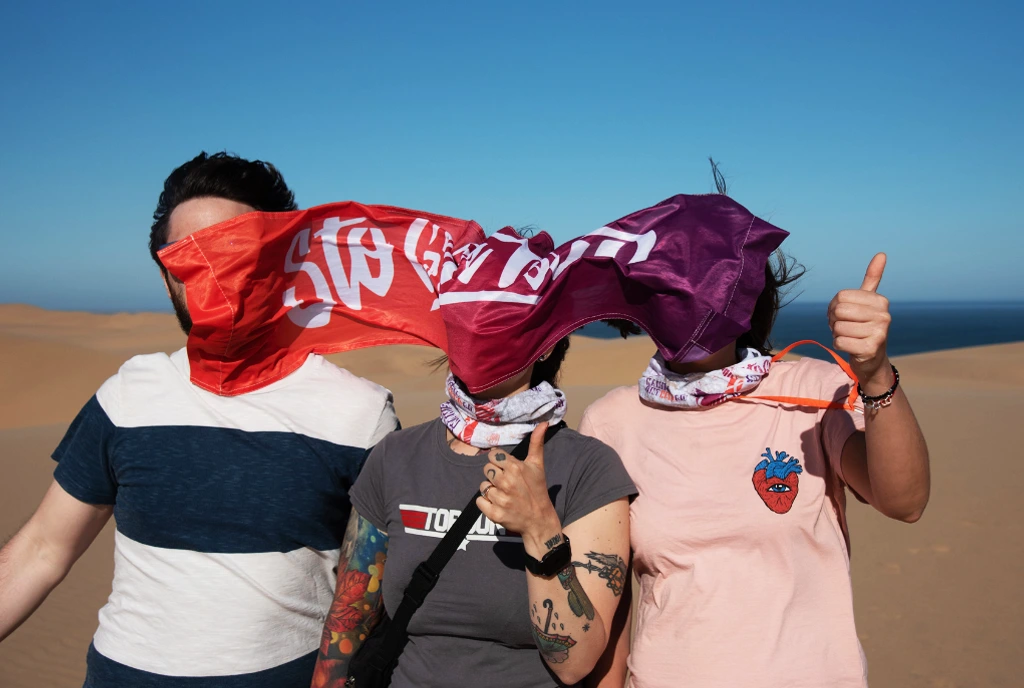 Three travelers on a dune by the sea, with the wind slapping the Sto Gran Tour banner in their faces